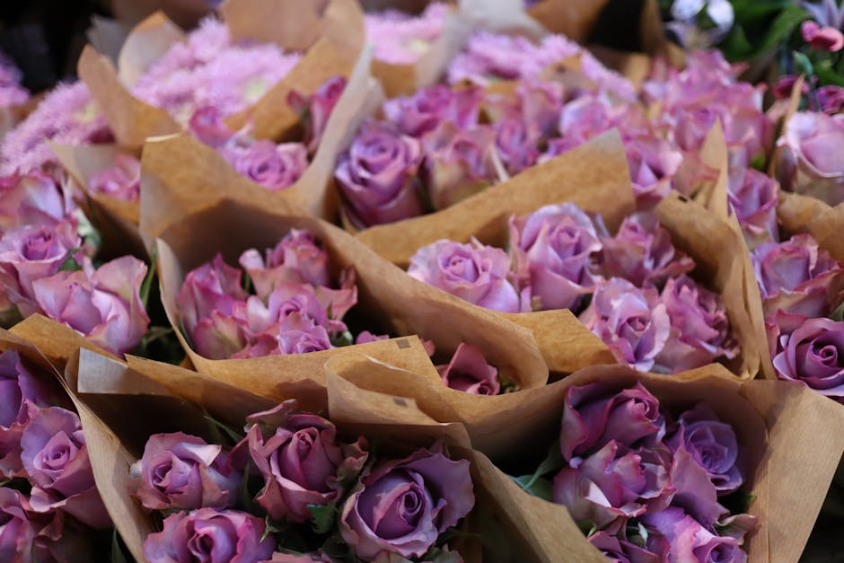 An abundant display of fresh purple roses arranged in small bouquets wrapped in light brown kraft paper, with some showcasing soft pinkish-purple petals and others in deeper violet shades. The bouquet