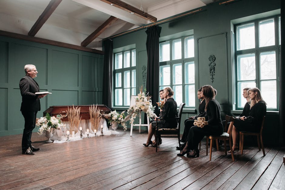 An indoor scene featuring a floral arrangement with predominantly white and cream-colored flowers, including roses and lilies, accented by dried grasses and greenery, placed on a white pedestal. The f