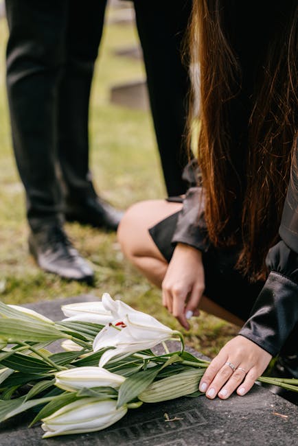 A person wearing a black jacket kneels on the ground, gently placing white lilies with green leaves on a memorial stone. The lilies are fresh, with delicate petals and long stems, arranged in a small 