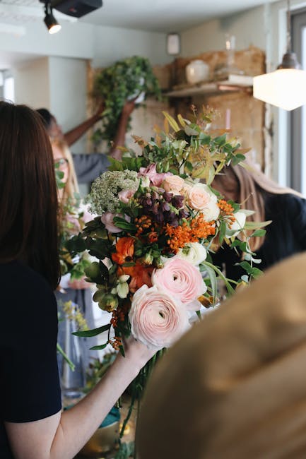A florist from company [COMPANY_NAME] is arranging a large, vibrant bouquet of fresh flowers indoors, with various blossoms including pink ranunculus, white hydrangeas, orange marigolds, and purple ac