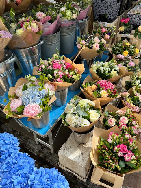 A vibrant display of fresh floral bouquets arranged in metal and cardboard containers, featuring a variety of flowers including pink roses, white carnations, light blue hydrangeas, and mixed greenery.
