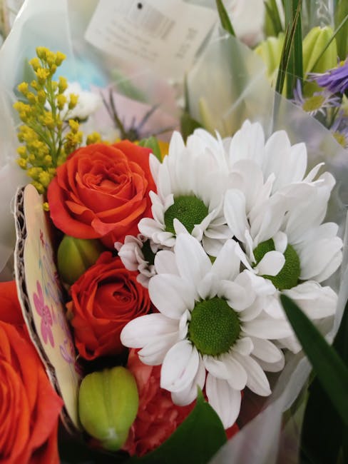 A close-up view of a vibrant floral bouquet featuring bright white daisies with green centers, paired with deep orange roses showcasing layered, velvety petals. The arrangement includes small yellow b