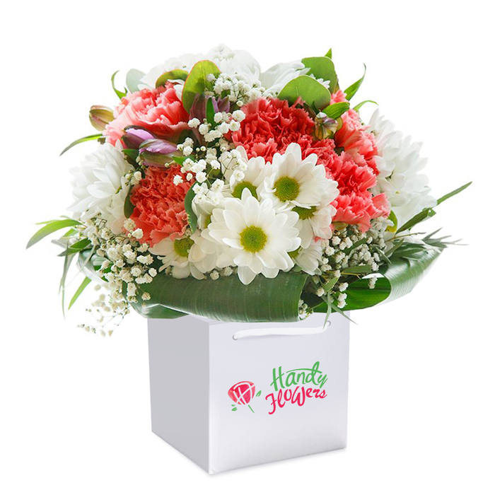 Within a crisp white square gift bag, lovingly arranged by a skilled Shooter's Hill florist, sits a bouquet that overflows with colour and texture-ready to uplift or comfort those who receive it. The arrangement is anchored by an abundance of soft coral and peach carnations, whose ruffled, layered petals exude an almost touchable softness, reminiscent of early spring blooms along the paths near Shooters Hill Golf Club. Between these, there are pure white daisies-each flower's faces bright with vivid, yellow-green cores-casting the kind of cheerful vibrance you'd find on a sunny day at Eaglesfield Park. Airy white gypsophila meanders throughout, its delicate clusters reminiscent of dewdrops caught in dawn light. Delicate, purple-hued buds, perhaps yet-to-bloom alstroemeria, gently peek from within the bouquet and lend a subtle complexity and anticipation to the mix. All of this is united by velvety, deep-green foliage and broad, shiny leaves, arcing around the base and sides to provide definition and a fresh, garden-like feel. Looming upright, the bouquet's rounded silhouette hints at luxury and care. The scene is heightened by the white bag's understated cord handles-suggesting easy, elegant transport, perhaps to a celebration at one of Shooter's Hill's beloved historic sites. Captured in a studio setting against a pure white background, this image brings together freshness, abundance, and the artistry of local flower delivery. It's an ideal representation of thoughtful floristry, making it perfect for special occasions or simply reminding loved ones in SE18 that they're cherished.