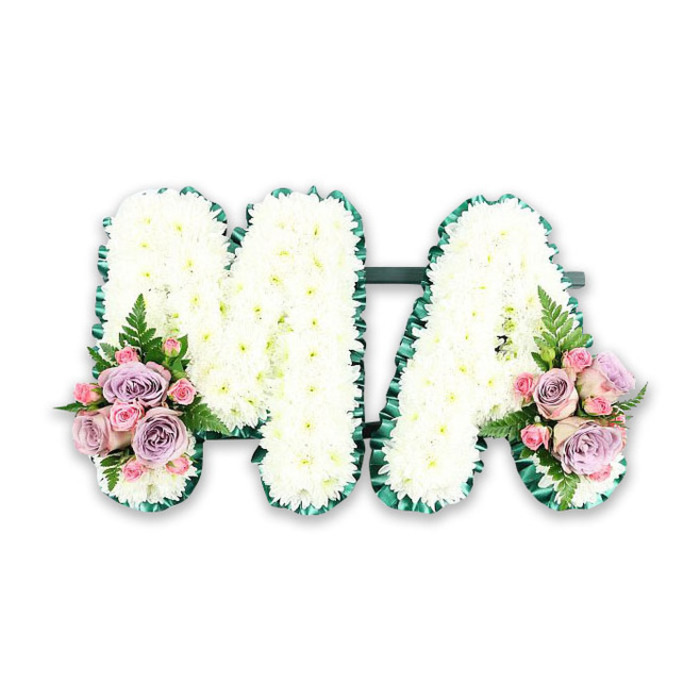 The image presents a serene funeral letter tribute, carefully spelling the word MA in bold, sculpted capitals. Each letter rests on a concealed frame, though only the luxurious blanket of flowers is visible. Dozens upon dozens of small, pristine white chrysanthemum blooms cover every surface, their rounded petals overlapping to create a dense, cloud-like texture. The white is clean and bright, evoking purity, peace, and the stillness that often surrounds a farewell ceremony. Tracing the outer contours of both letters is a pleated band of dark, emerald green ribbon or foliage, carefully folded and pinned so that it follows every straight line and angle of the M, then curves smoothly around the arch and crossbar of the A. This deep green edging not only defines the shape but also adds a sense of craftsmanship and intention, much like the thoughtful floristry often requested for services in and around Shooter's Hill. At the base of the left stem of the M and the right stem of the A, symmetrical clusters of coloured flowers soften the starkness of white. Here, pale pink roses mingle with cool lilac and lavender roses, while miniature pink spray roses nestle between them like little jewels. Wisps of fern fronds and glossy green leaves peek out, adding texture and a natural, garden-inspired feel. The arrangement is lit evenly, ensuring that every petal and fold can be distinguished, making it suitable as a catalogue-style product image for funeral flower delivery in the Shooter's Hill area. Emotionally, it speaks of a gentle, heartfelt farewell to a mother or mama, a simple yet profound way to spell out love and remembrance in flowers.